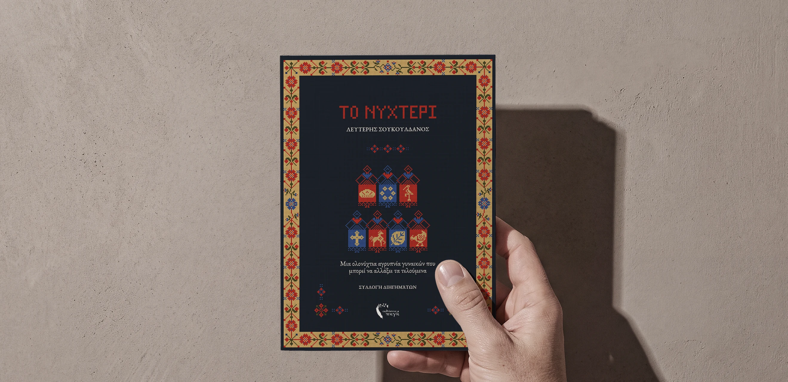 The finished book held against a pale wall - ΤΟ ΝΥΧΤΕΡΙ in red on navy, inside a woven folk border, with seven stitched panels set three over four down the middle