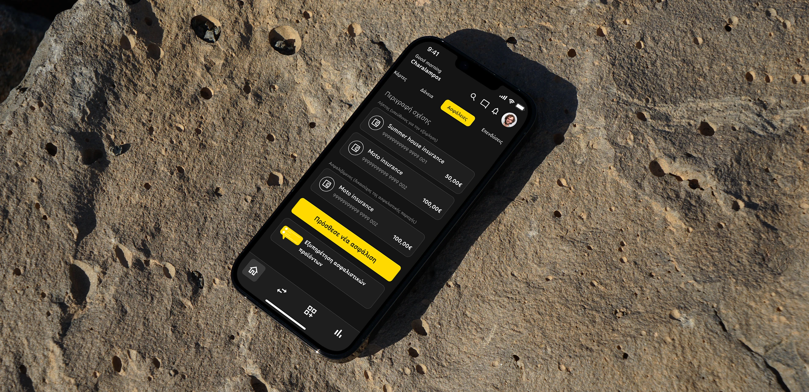A phone lying on sun-lit rock, showing the insurance section of the banking app in dark mode with a yellow button